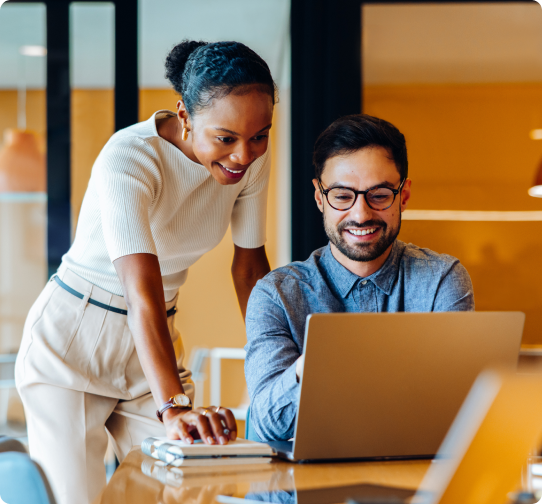 A professional man and woman in an office space work on a laptop together.

