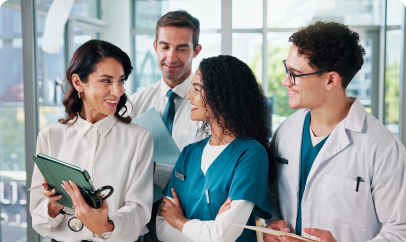 A group of healthcare professionals in scrubs and white coats collaborate and discuss something on a tablet.